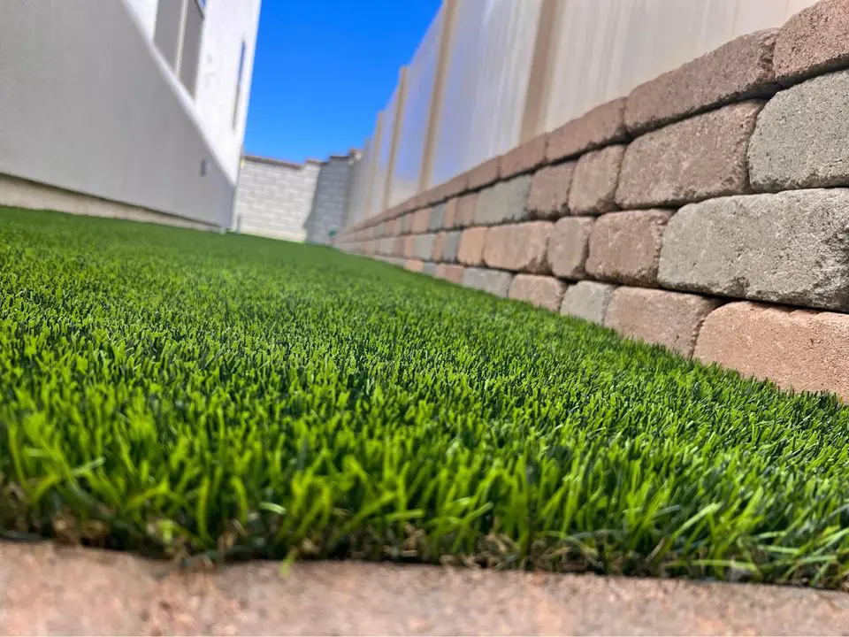 Close-up of a narrow, neatly trimmed green lawn bordered by a low stone retaining wall on the right and the side of a white building on the left. The wall and building create a corridor effect, showcasing artificial grass by Scottsdale Turf Pros under a clear blue sky.