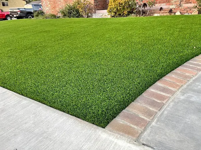 A well-manicured green lawn, featuring high-quality artificial grass, is bordered by a brick edge adjacent to a concrete sidewalk. In the background, there is a red brick building with shrubs and other plants. This beautiful Cave Creek landscape exemplifies meticulous attention to detail.