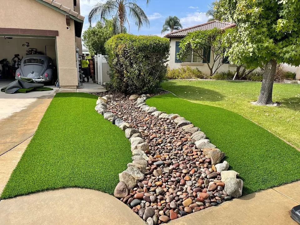 A well-maintained front yard with artificial grass, featuring a dry riverbed made of pebbles and bordered by large rocks. The area includes a tree, trimmed bushes, and a driveway leading to a garage with an old Volkswagen Beetle. Scottsdale Turf Pros completed the pet turf installation. Another house is visible in the background.