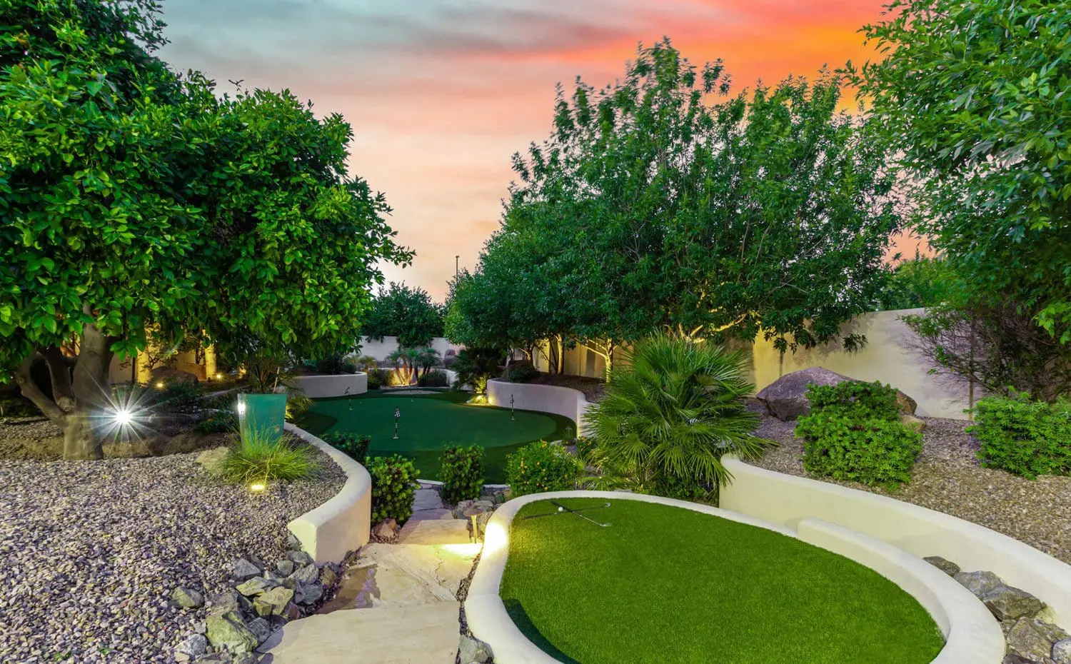 A beautifully landscaped backyard features a putting green surrounded by trees and shrubs. Curved white retaining walls enclose the area, and small lights illuminate the space at dusk, creating a serene and inviting atmosphere—courtesy of Scottsdale Turf Pros in Scottsdale AZ.