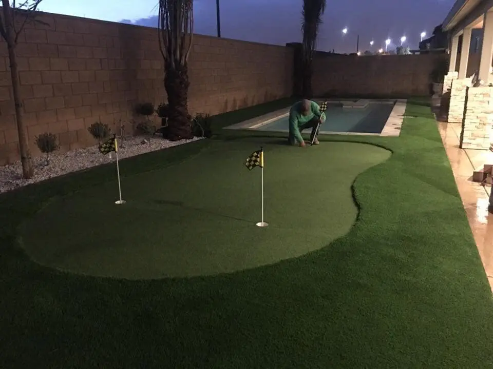 A person is kneeling and working on a putting green with artificial sports turf in a backyard. The putting green has two holes with checkered flags, surrounded by vibrant faux grass. This Scottsdale, AZ backyard also features a narrow swimming pool adjacent to a wall. It is dusk or nighttime.