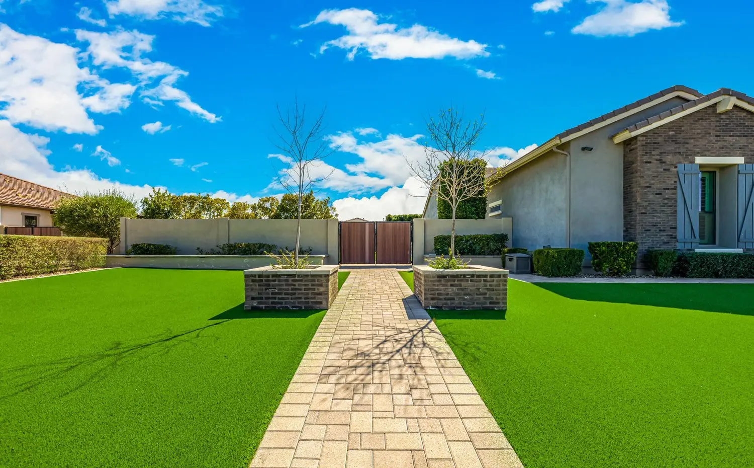 A well-manicured backyard with artificial green grass, courtesy of Scottsdale Turf Pros, features a paved stone walkway leading to a wooden gate. Alongside the walkway are raised brick planters with young trees, and on the right, there's part of a house with a brick and stucco exterior. Blue sky with clouds.