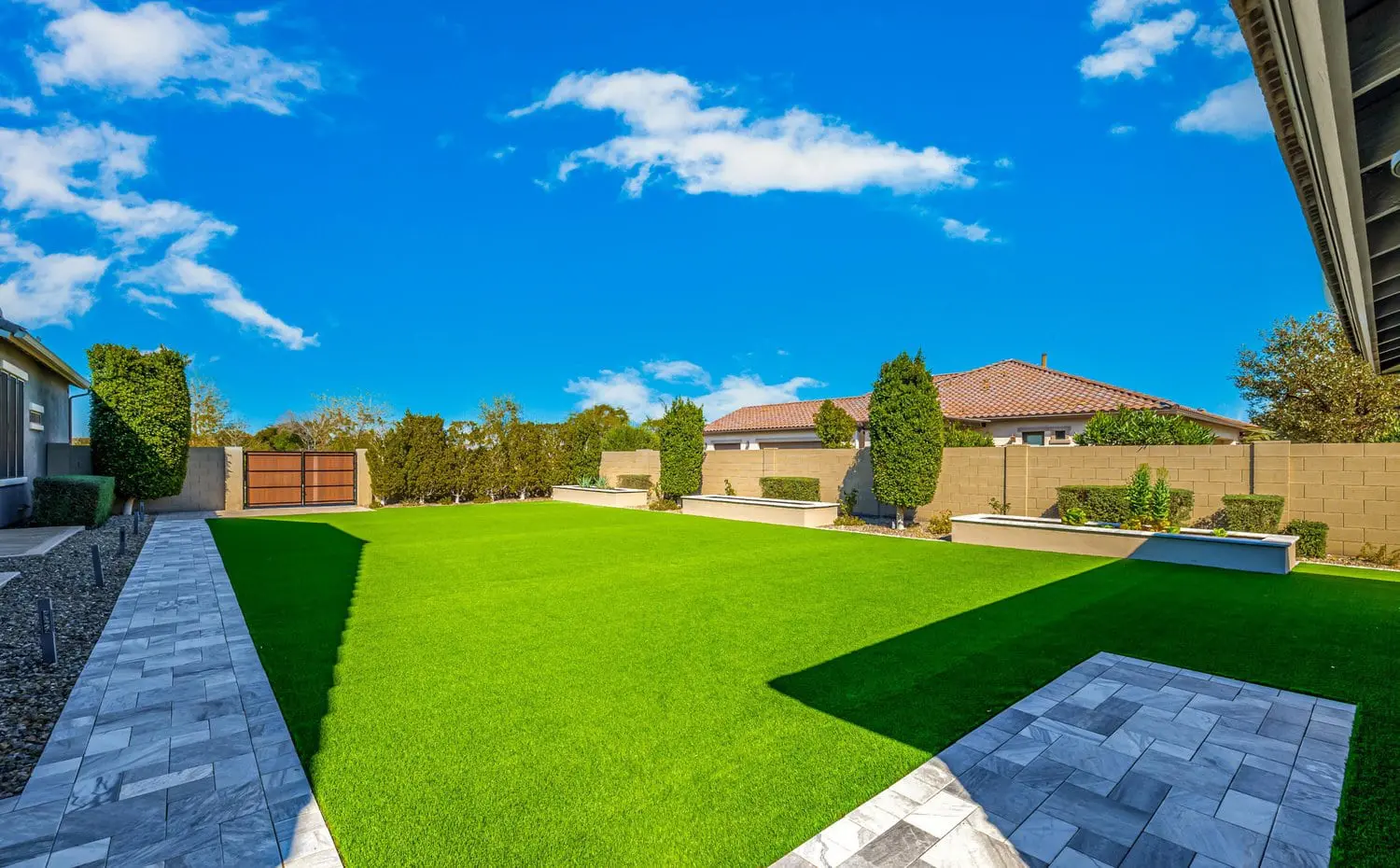 A well-maintained backyard with a large lawn of bright green artificial grass, expertly installed by Scottsdale Turf Pros, framed by stone pathways. The yard has a few tall shrubs, some behind stone planters, and is surrounded by a stone wall. In the background, houses with orange tiled roofs bask under the blue Phoenix AZ sky.