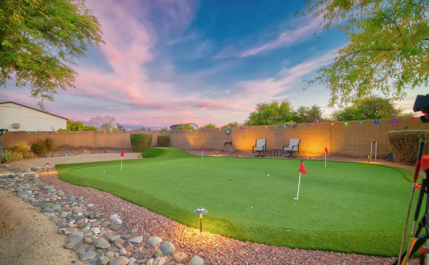 A backyard putting green is shown at sunset, with a vibrant sky of pink and blue hues. The artificial turf installation features multiple holes with small red flags. It is bordered by a stone path and trees. Two chairs and a low wall with decorative lights complete the picturesque setting in Scottsdale AZ.