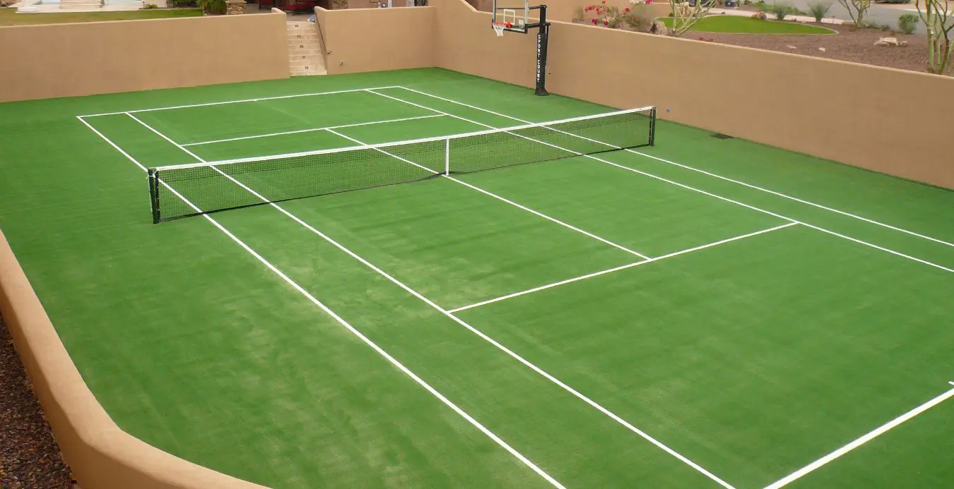 A backyard putting green is shown at sunset, with a vibrant sky of pink and blue hues. The artificial turf installation features multiple holes with small red flags. It is bordered by a stone path and trees. Two chairs and a low wall with decorative lights complete the picturesque setting in Scottsdale AZ.