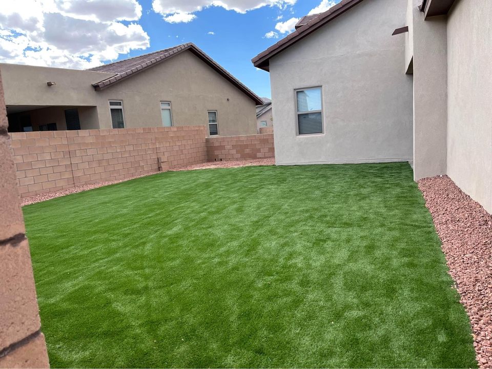 A backyard with artificial green grass from Scottsdale Turf Pros, bordered by reddish-brown gravel. The yard is enclosed by beige stucco house walls on three sides and a stone block wall on the fourth side. Overhead, a blue sky with scattered clouds is visible in Scottsdale, AZ.