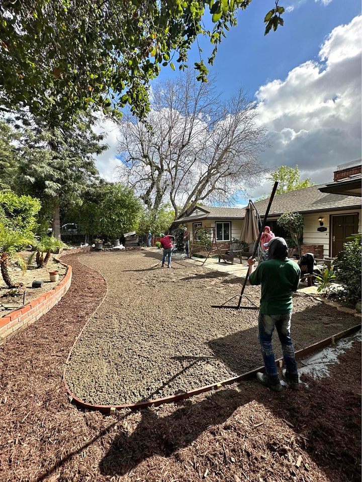 Workers are leveling a layer of gravel in a backyard, preparing for an artificial grass installation. They are using long tools to smooth out the area. Surrounding the yard are plants, a tree with bare branches, and a house with patio furniture under a large umbrella.