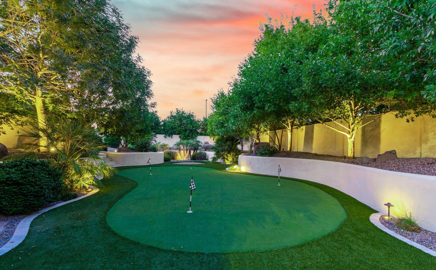 A backyard putting green at dusk, featuring artificial grass installation, is surrounded by well-manicured trees and soft lighting. The sky shows shades of pink and orange, creating a serene atmosphere. Small checkered flags mark the holes on this maintenance-free lawn.