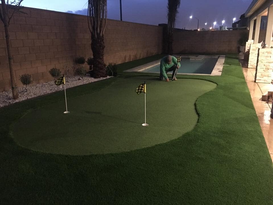 A person is kneeling and working on a putting green with artificial sports turf in a backyard. The putting green has two holes with checkered flags, surrounded by vibrant faux grass. This Scottsdale, AZ backyard also features a narrow swimming pool adjacent to a wall. It is dusk or nighttime.