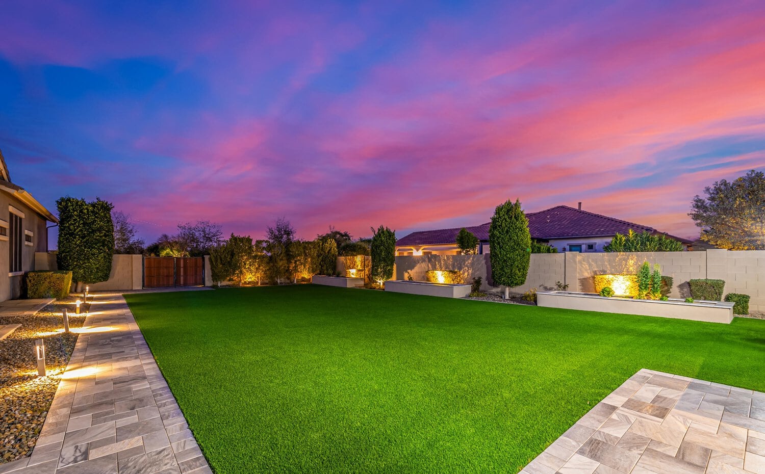 A beautifully landscaped backyard at sunset, featuring a lush green Tempe lawn installed by Scottsdale Turf Pros, with stone pathways and modern rectangular planters holding trees and bushes. The sky displays vibrant hues of pink and purple, casting a serene ambiance over the scene.