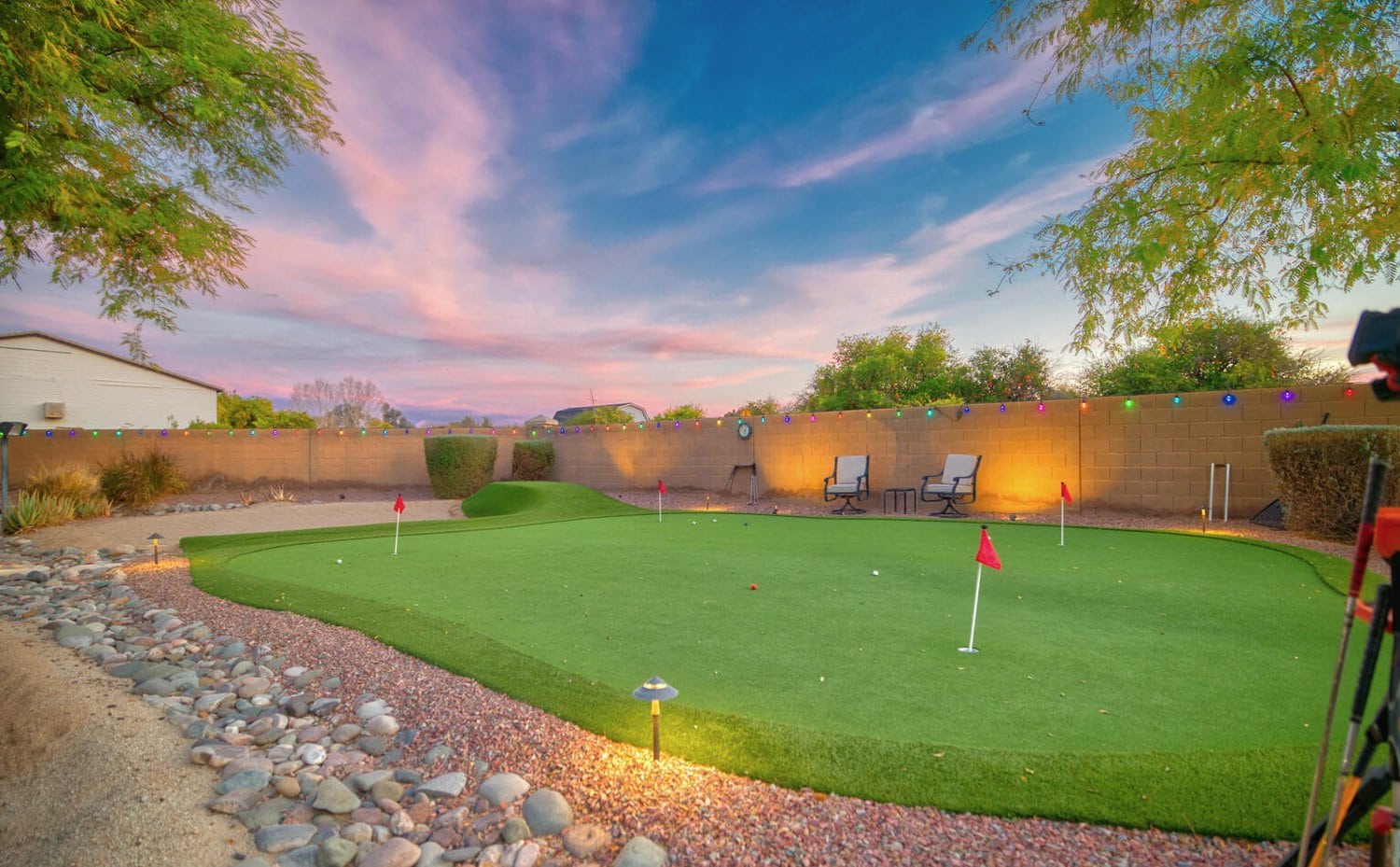 A backyard features a well-manicured artificial putting green with red flags and nearby landscaping. Two chairs sit on pavers against a cinder block wall adorned with festive string lights. The sky is vibrant with pink and purple hues, suggesting a sunset or sunrise.