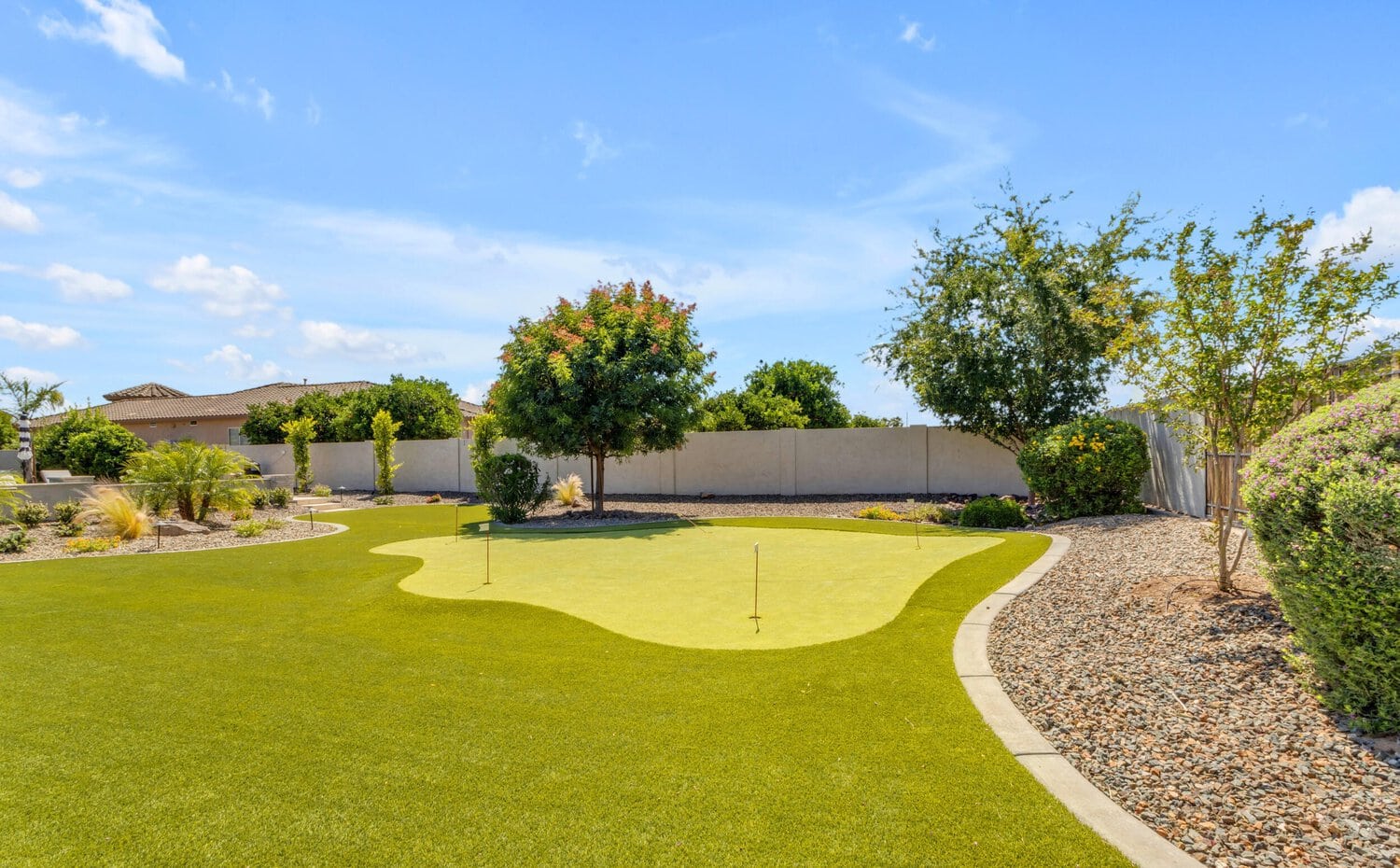 A well-manicured backyard with a putting green featuring two flags, surrounded by lush greenery, shrubs, pebbles, and a stone pathway. Trees and a white fence border the area, with a clear blue sky overhead—expertly crafted by Scottsdale Turf Pros using premium artificial grass for durability and beauty.