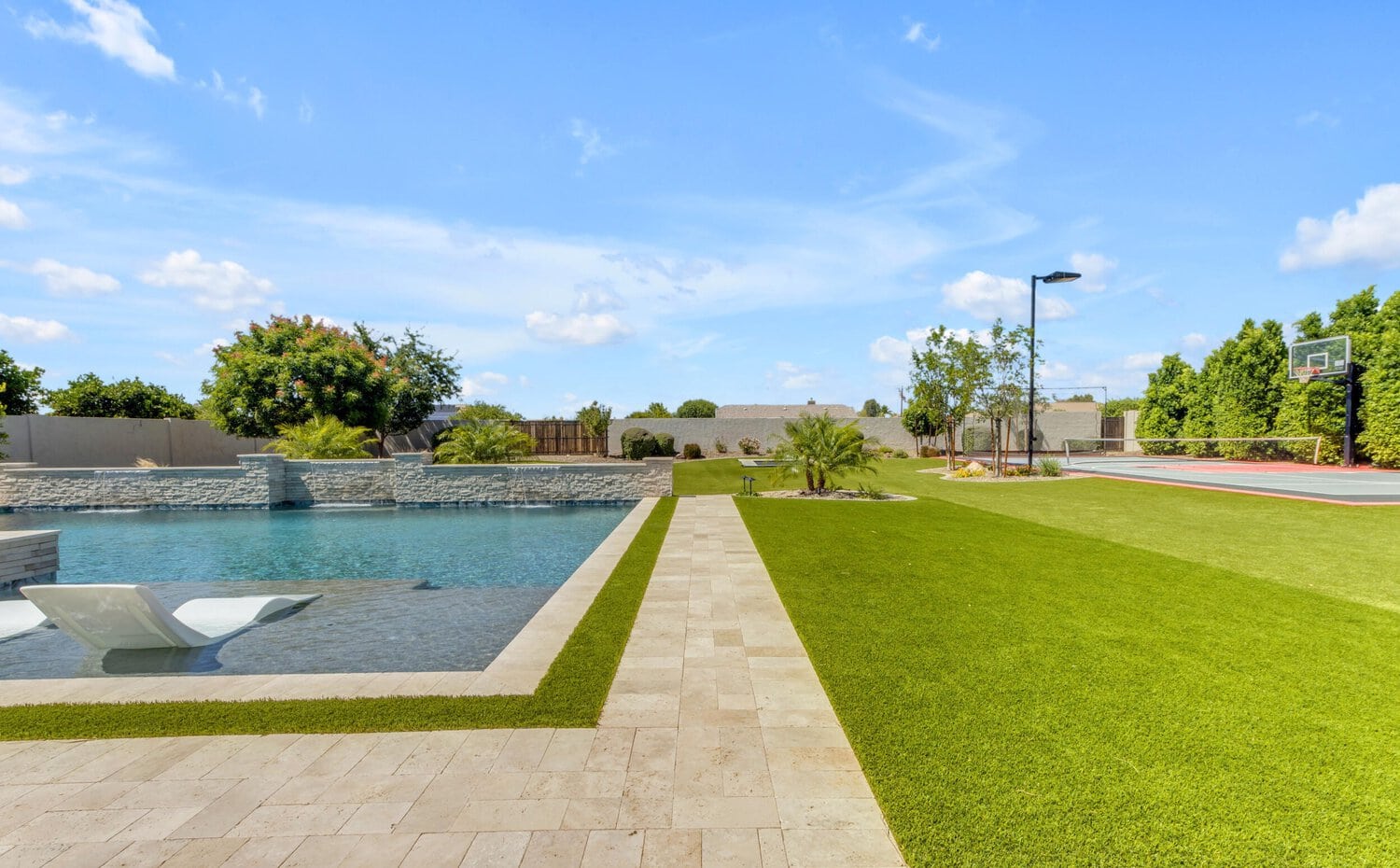 A luxurious backyard features a modern swimming pool with in-pool lounge chairs and a grassy area leading to a basketball court. The scene is bright and sunny, with a clear blue sky and neatly manicured trees and shrubs surrounding the space, all expertly designed by Scottsdale Turf Pros.