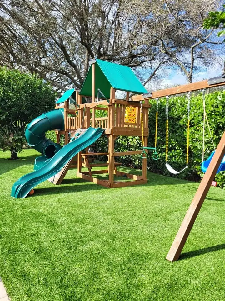 A playground with fresh, green artificial turf installed under it. The playground has dark green slides, 3 swings, a climbing frame, and more.