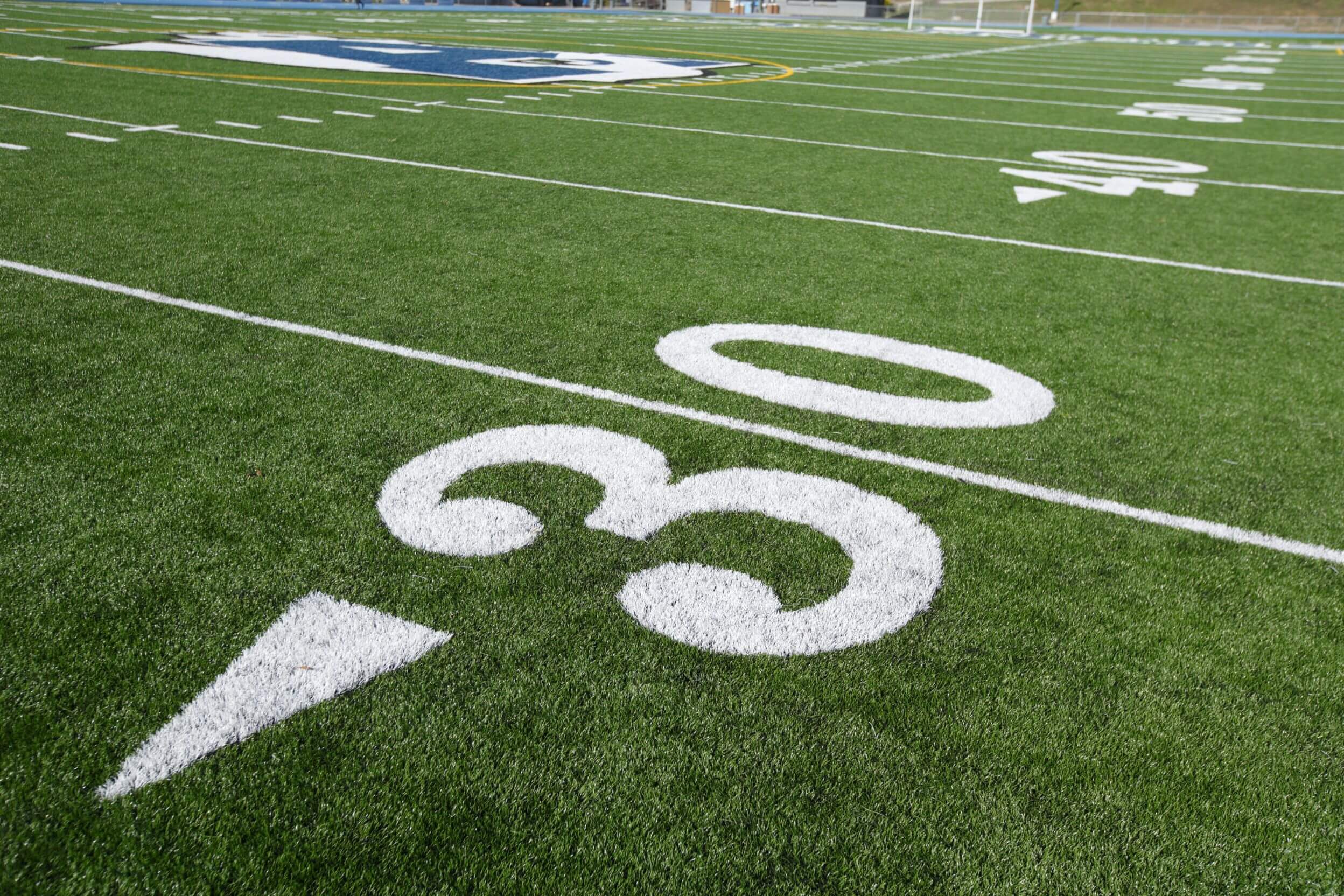 A close-up of an American football field, focusing on the number 30 yard line marker. The bright green artificial turf, installed by Scottsdale Turf Pros, boasts crisp white yard lines and numbers. Part of a team logo is visible in the background on the field surface.