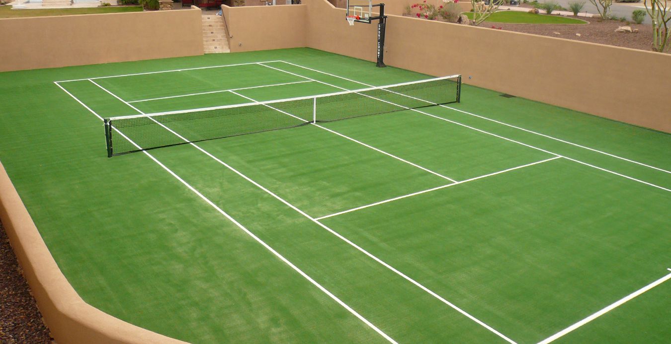 A neatly maintained multi-use sports court in Phoenix, AZ combines tennis and basketball in an enclosed area with tan walls. The court features a green surface with white lines, a net in the center for tennis, and a basketball hoop on one end. Stairs lead up to the Scottsdale Turf Pros artificial grass installation.