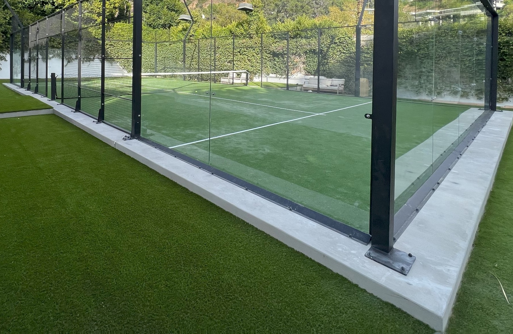 A person is kneeling and working on a putting green with artificial sports turf in a backyard. The putting green has two holes with checkered flags, surrounded by vibrant faux grass. This Scottsdale, AZ backyard also features a narrow swimming pool adjacent to a wall. It is dusk or nighttime.